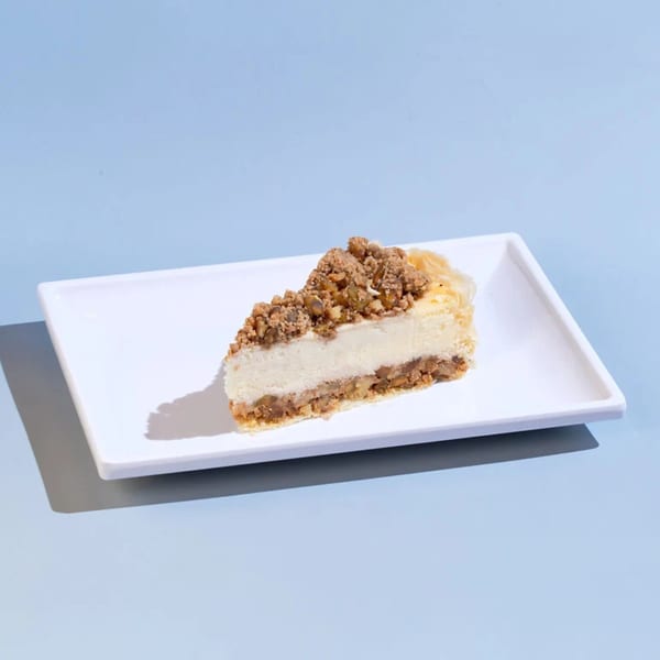 A slice of cheesecake with a crumbled topping and a flaky crust on a white rectangular plate.
