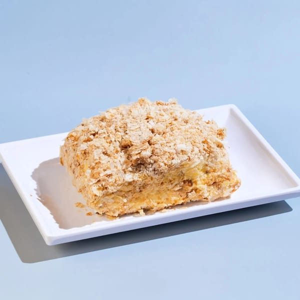 A slice of Efi's Gyro's Greek Napoleon cake, topped with flaky pastry crumbs, on a white plate.