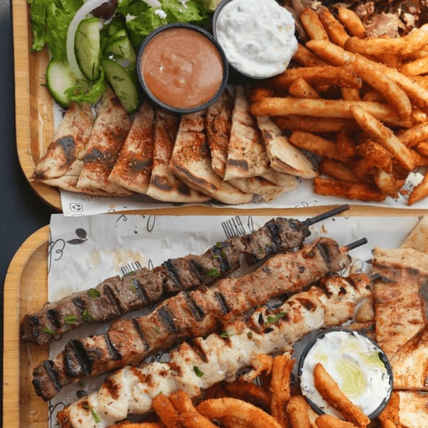 Two wooden trays filled with Greek food, including skewers, pita, fries, salad, and dips.