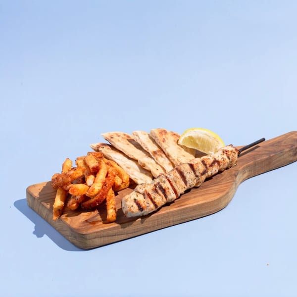 Efi's Gyro chicken souvlaki skewer, seasoned fries, pita bread, and lemon wedge on a wooden board.