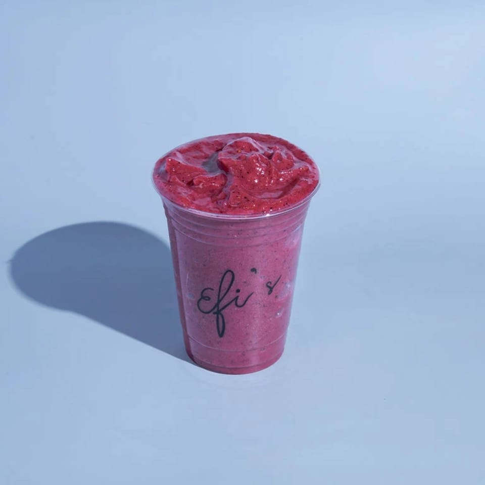 A vibrant red berry smoothie in a clear cup with "Efi's" logo, casting a shadow on a light blue surface.