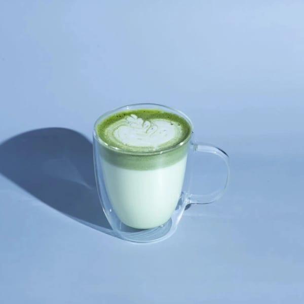 A clear glass mug with a green matcha latte and white foam art on a light blue background.